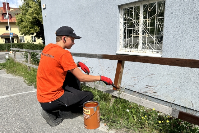 En ung person i orange t-shirt och svart keps sitter på huk och målar ett staket invid en parkering