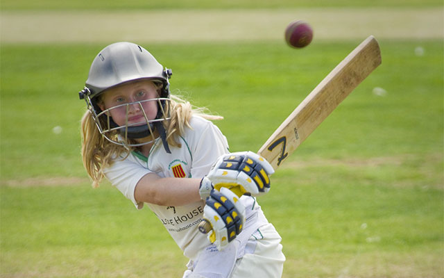 Ett barn med långt ljust hår klädd i cricket kläder slår en cricketboll