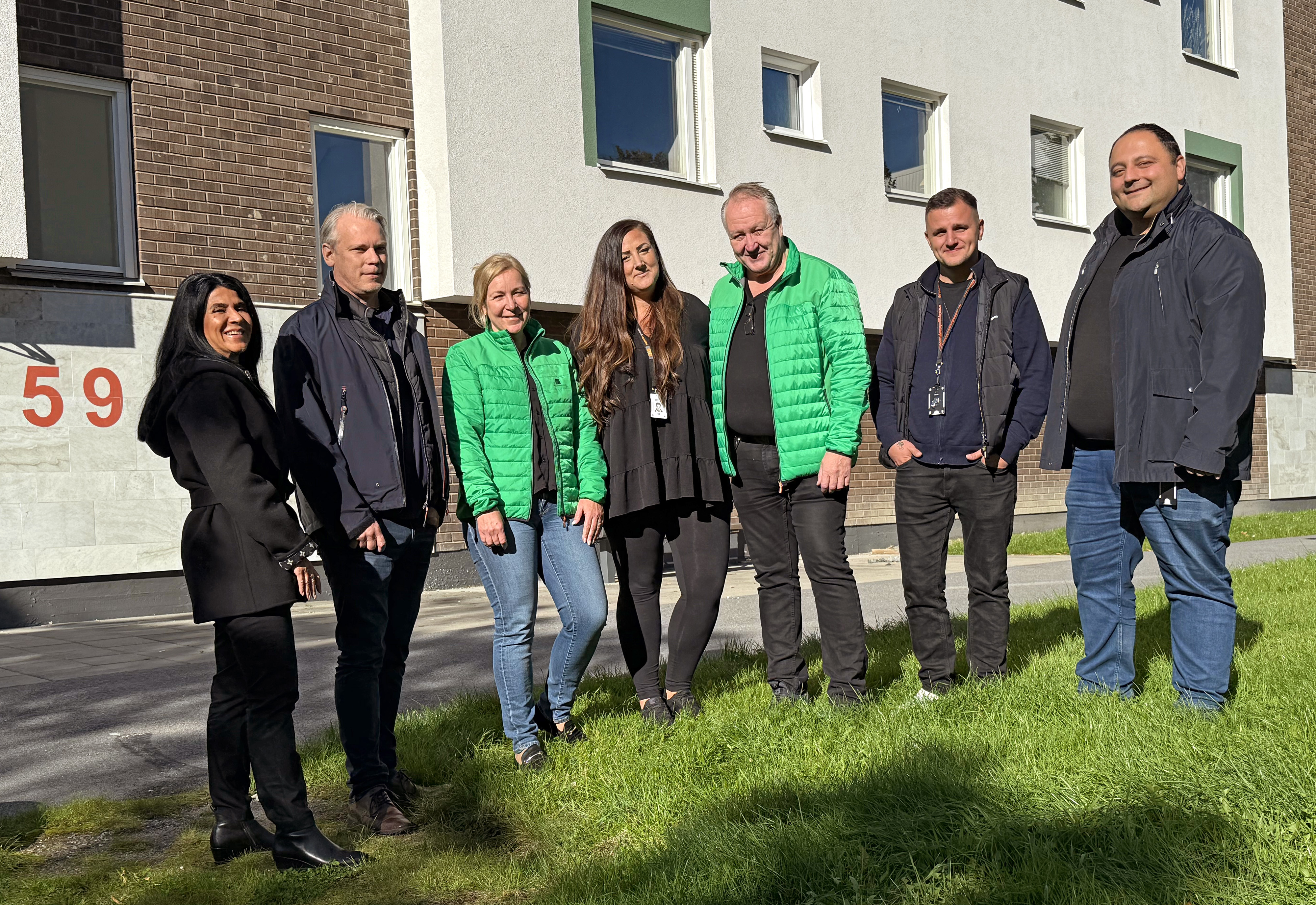 Sju personer står på en gräsmatta framför ett flerbostadshus.