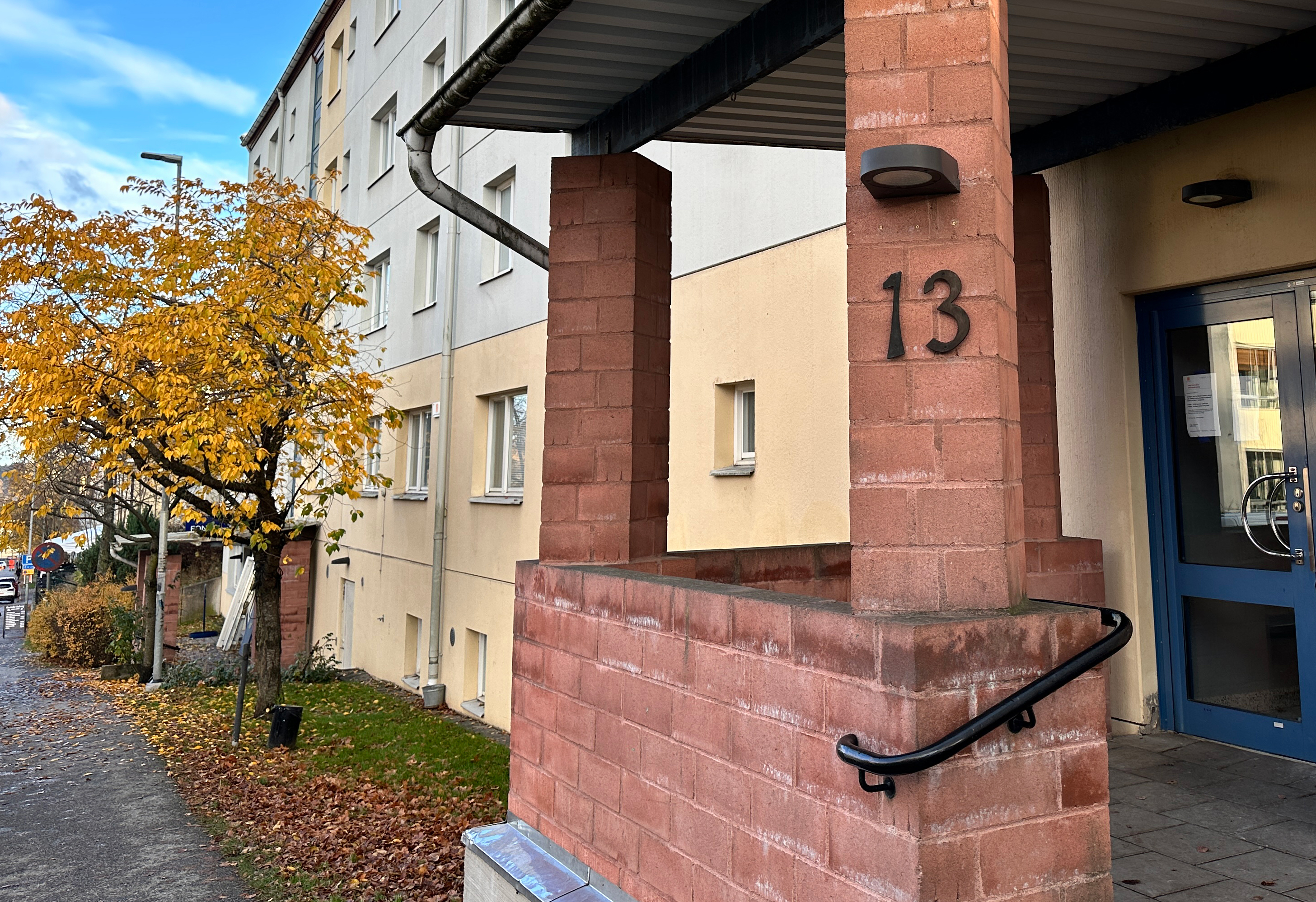 Entré i rött tegel till ett flerbostadshus med ljus putsad fasad. I bakgrunden ett träd med höstlöv.