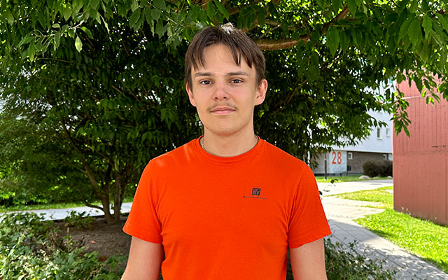 En kille i orange t-shirt står framför ett grönskande träd.