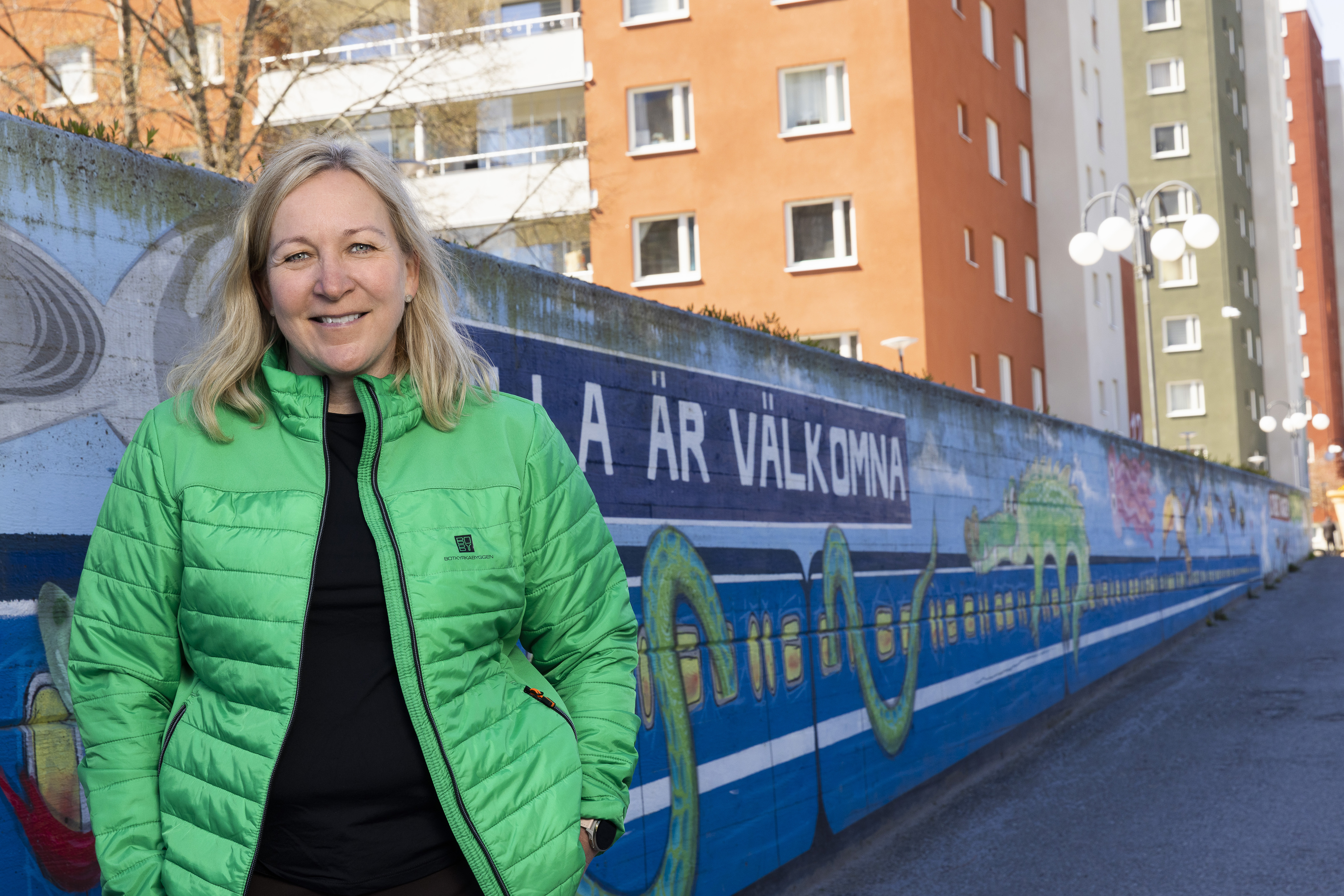 Kvinna i grön jacka framför målad mur och bostadshus.