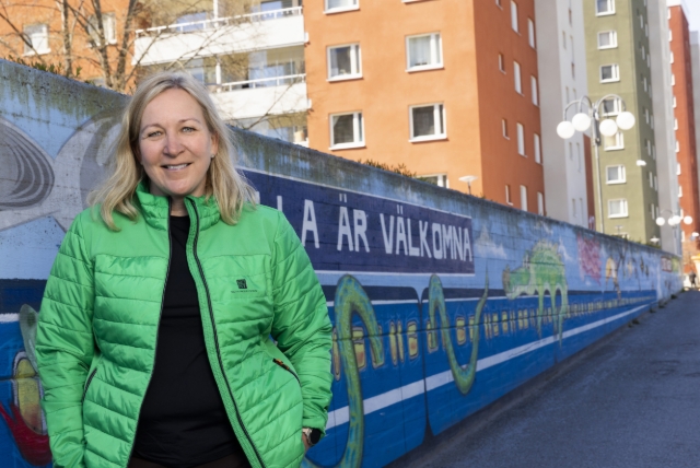 Kvinna i grön jacka framför målad mur och bostadshus.
