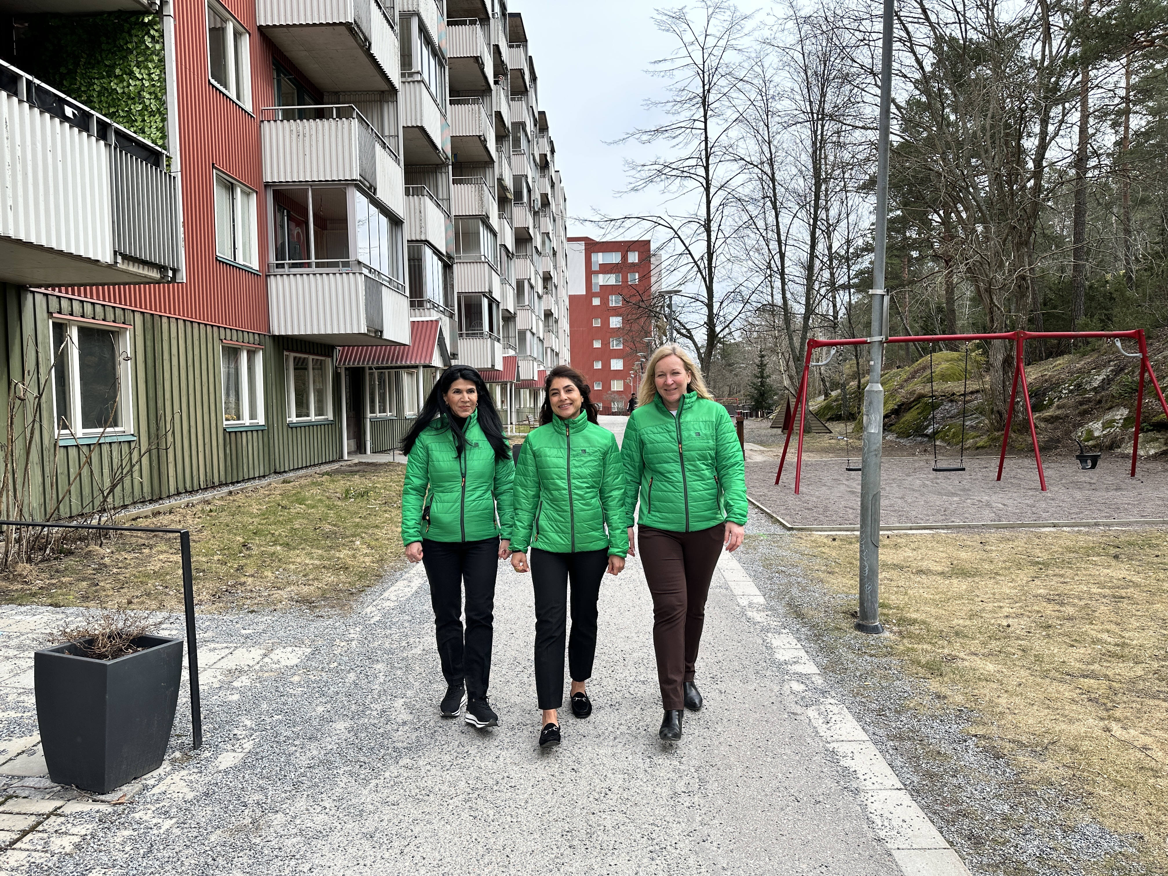 Tre kvinnor i gröna jackor går på en gångväg framför ett hus.