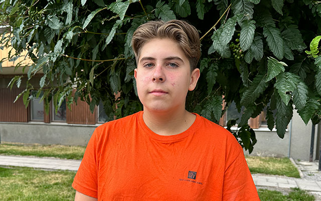 En kille i orange t-shirt står framför ett grönskande träd.