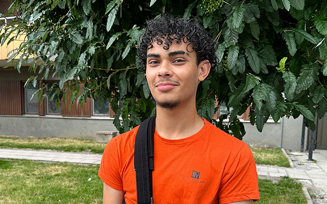En kille i orange t-shirt står framför ett grönskande träd.