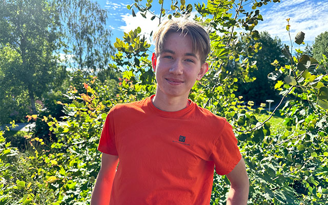 En kille i orange t-shirt står framför ett grönskande träd.