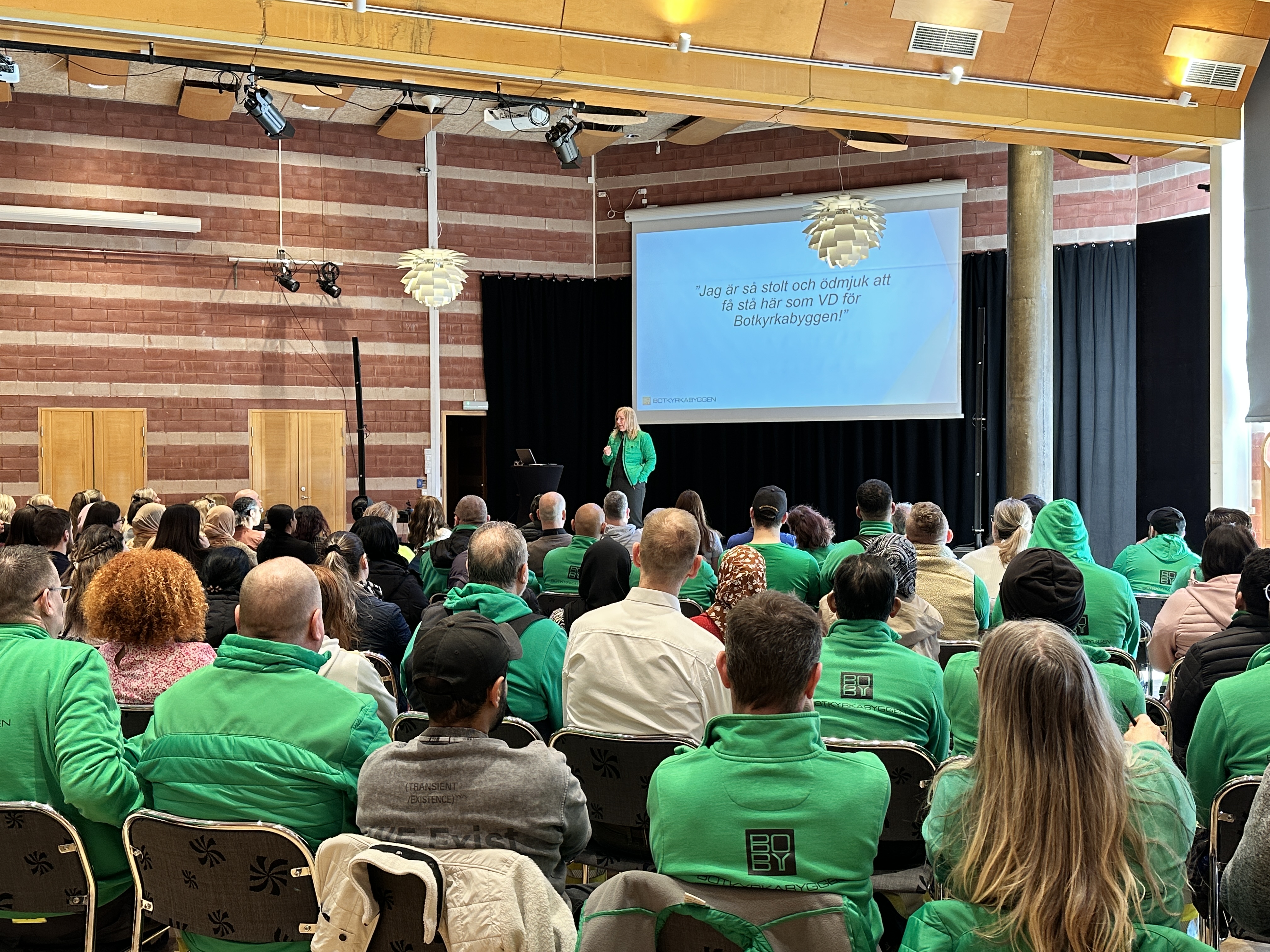 En grupp människor i gröna kläder sitter och lyssnar på en kvinna som står på en scen och pratar i 