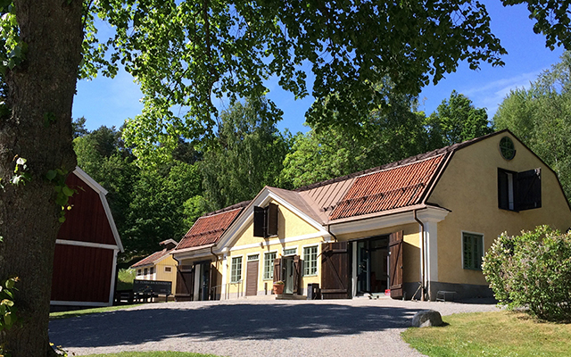 Ett stort gult hus med en grusplan framför. Blå himmel och grönskande träd runt om.