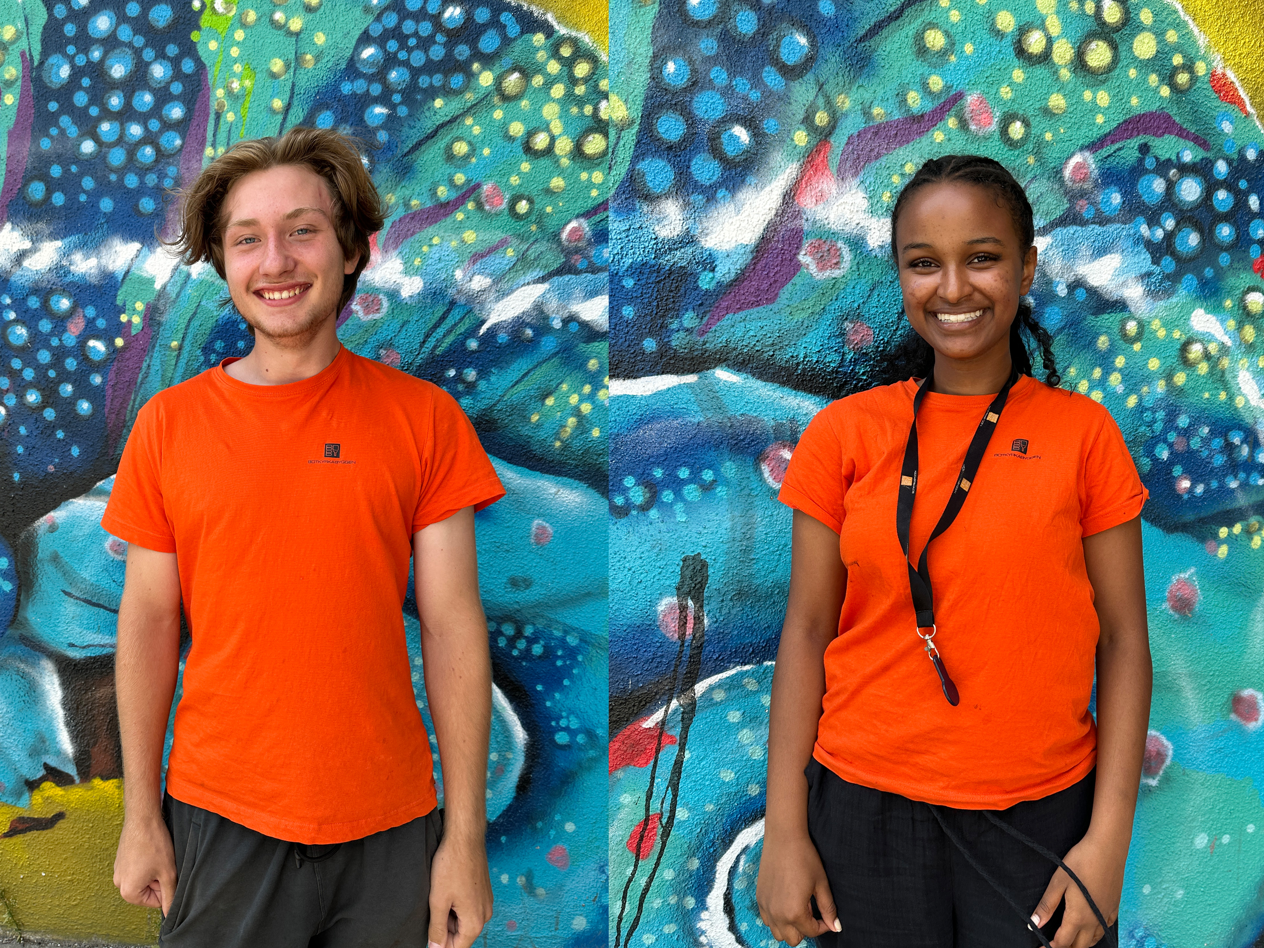En kille och en tjej i orange t-shirts står framför en färgglad väggmålning i blå och gröna toner